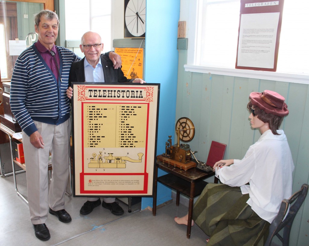 Sune Kvist tar emot gåvan “Telehistoria” av SI9AMs ordförande SM3FJF/Jörgen Planschen “Telehistoria” placerades därefter i fönstret ovanför kvinnas som sitter och telegraferar. Foto: SM3HKN Kenneth